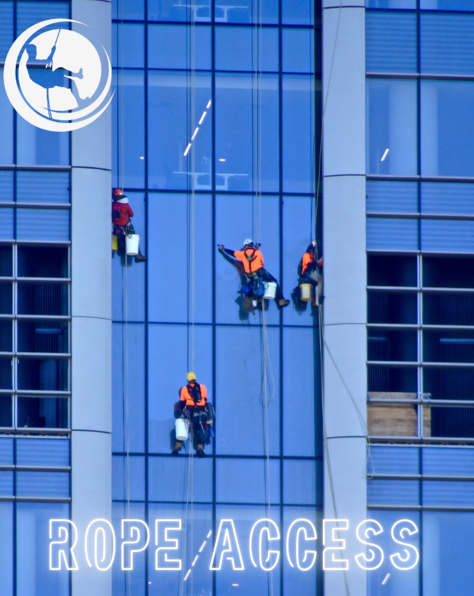 Window washers using ropes to clean a tall building's glass facade. Orange vests, blue sky reflected.