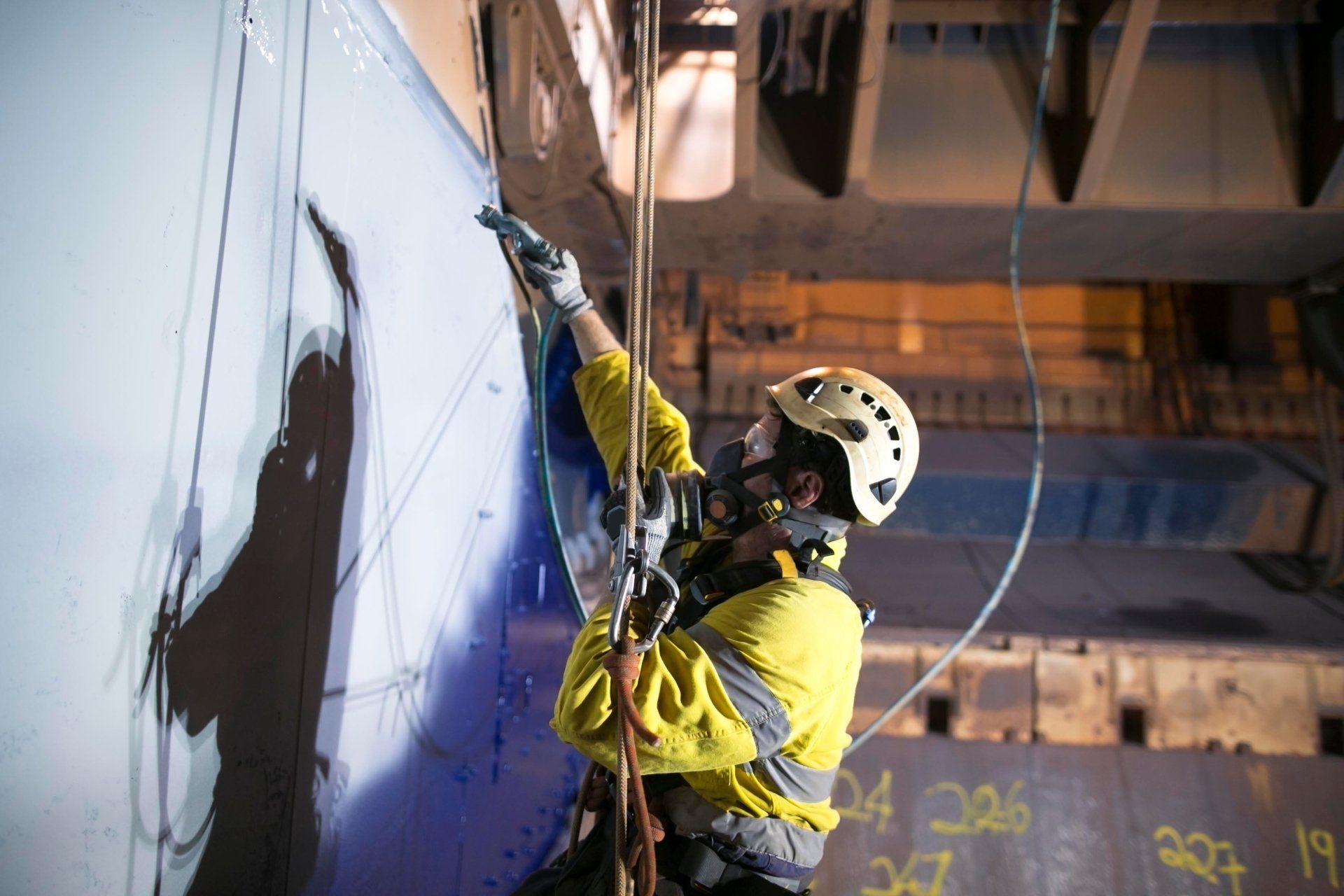 a man wearing a helmet and a face mask while dangling and painting a wall