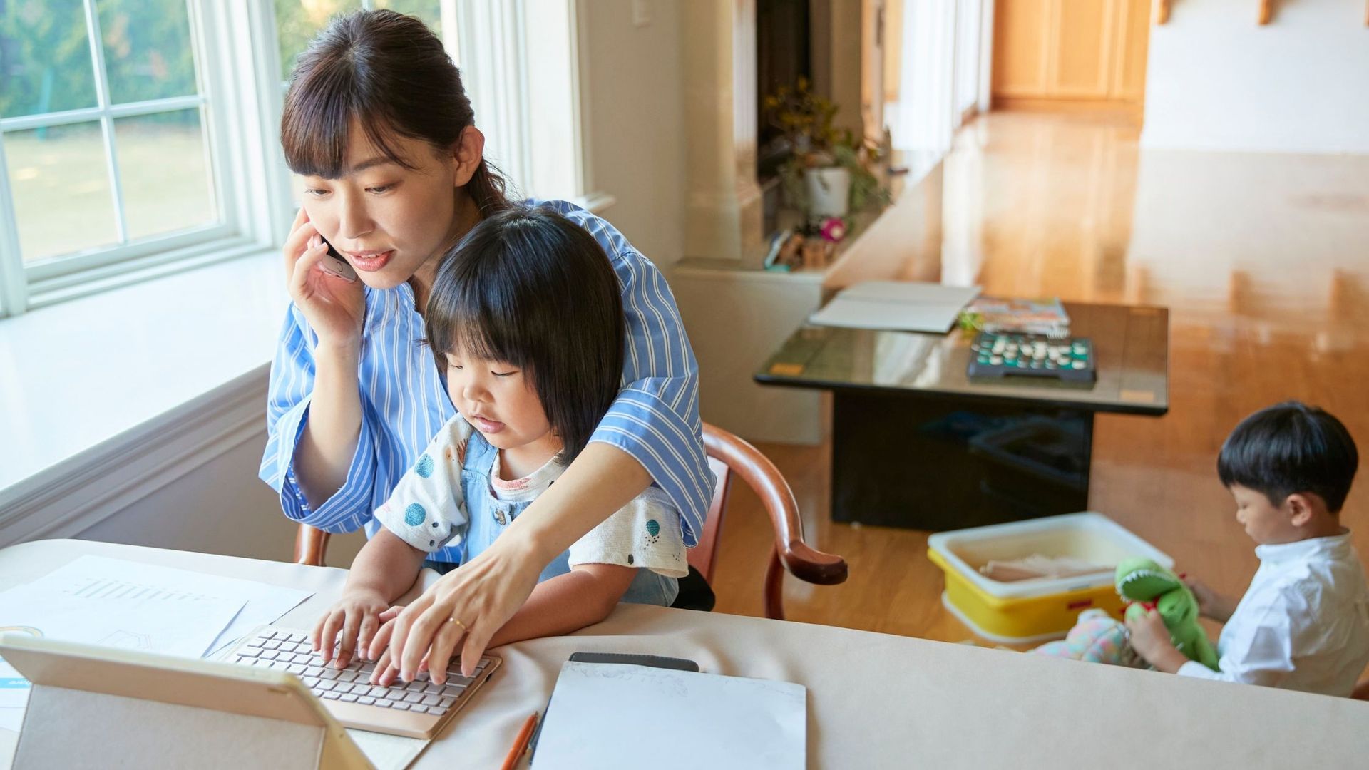 Woman on phone, working on laptop with child, other child playing. Bright, home setting.