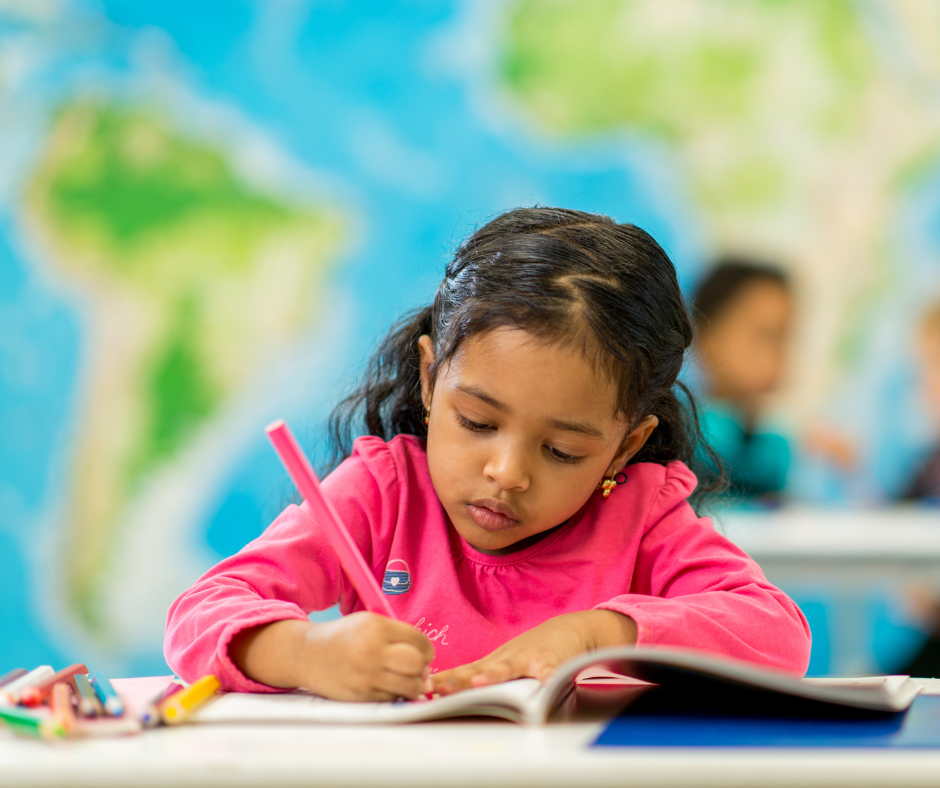 A little girl is sitting at a desk in a classroom writing in a notebook.