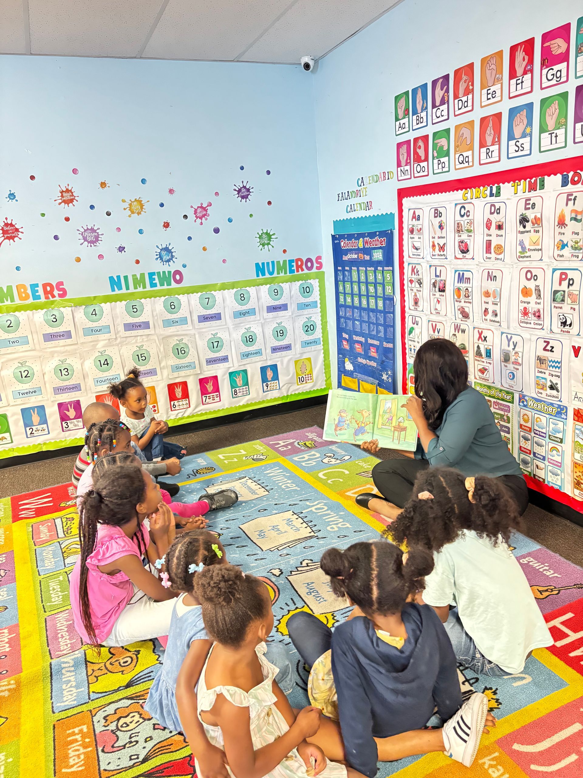 A group of children are sitting on the floor in a classroom listening to a teacher read a book.