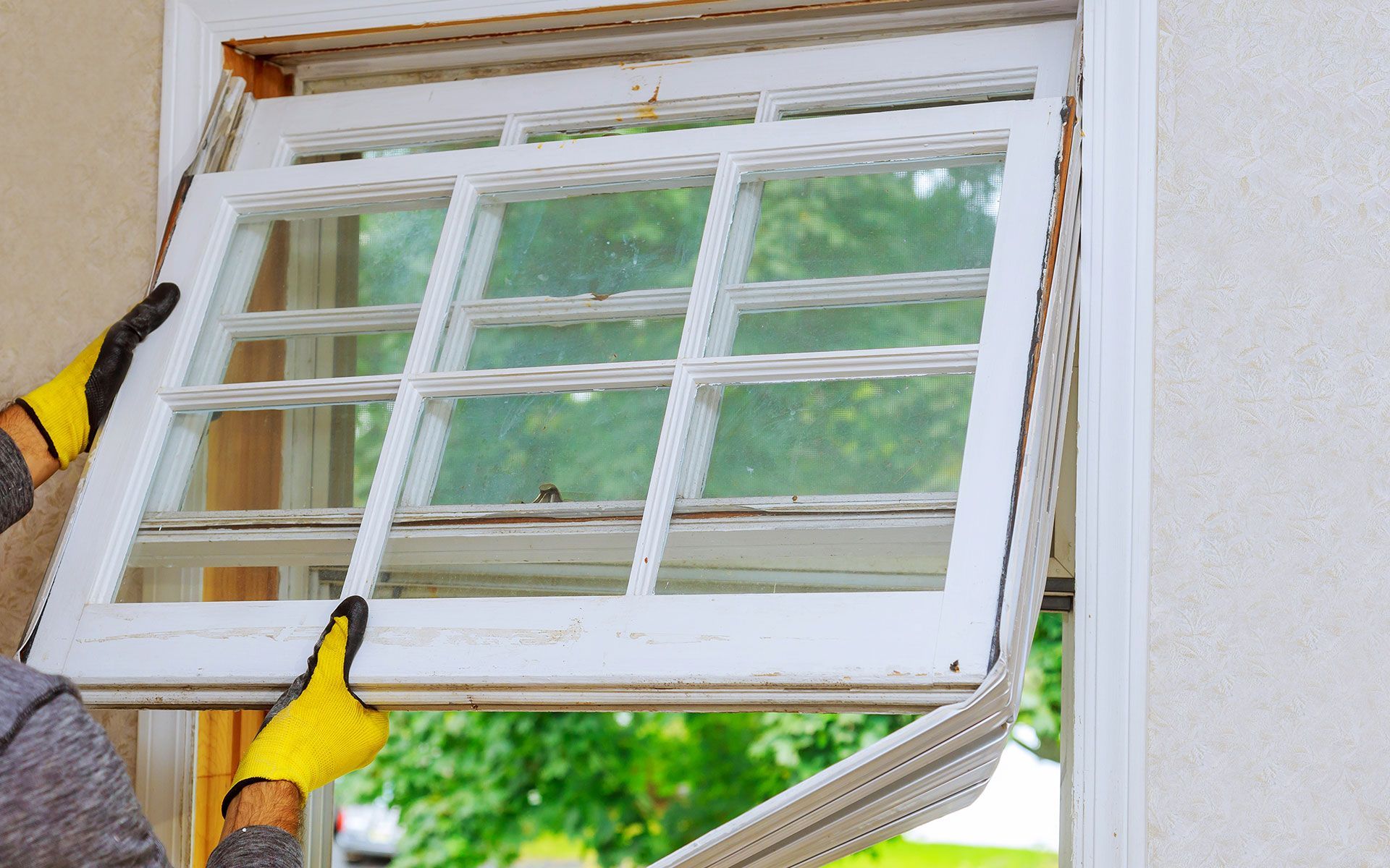 Man on a ladder installing something on a wooden window frame with a view of trees outside.
