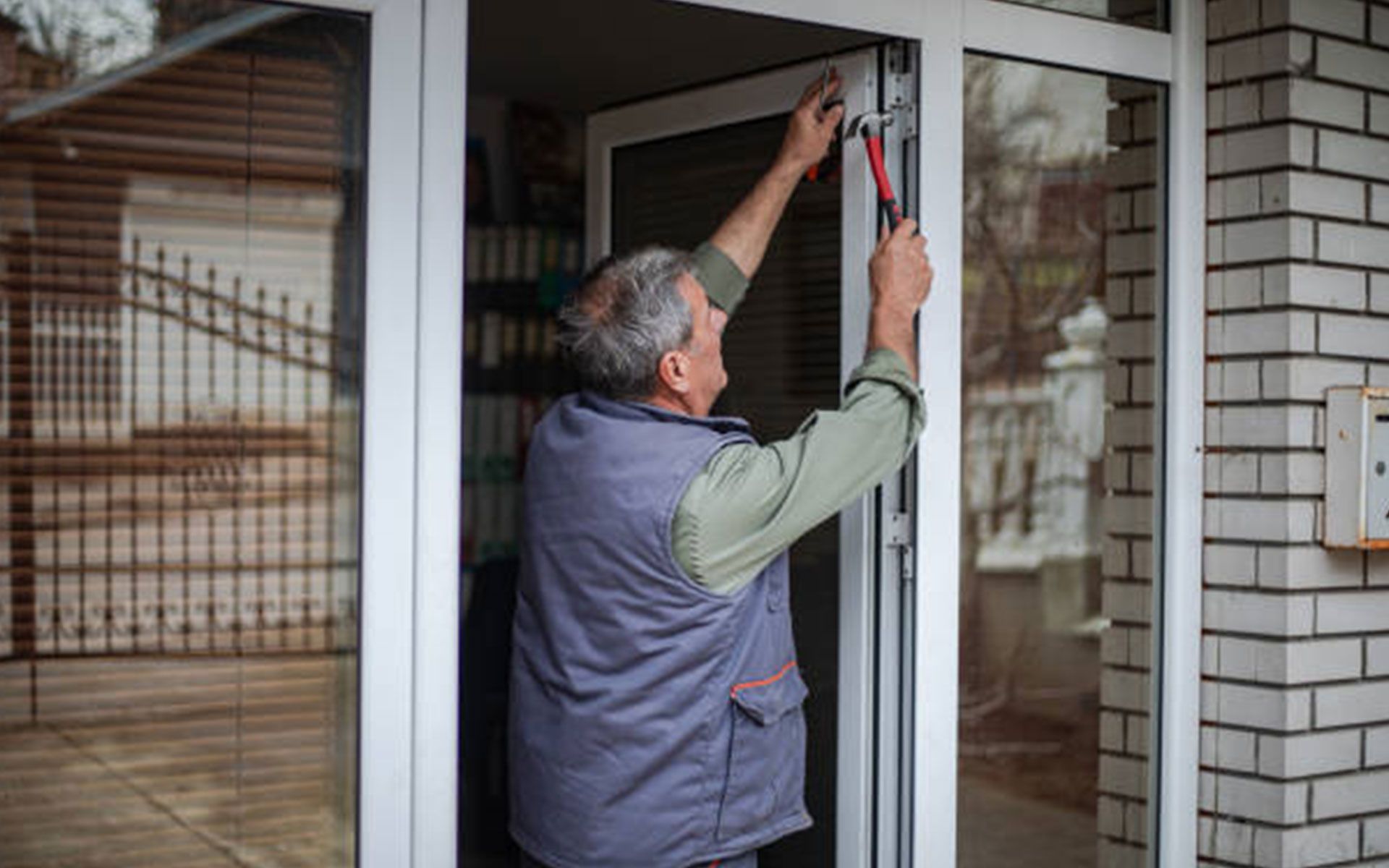 Man repairing a window frame with pliers. Standing outside near a brick building, gray vest.