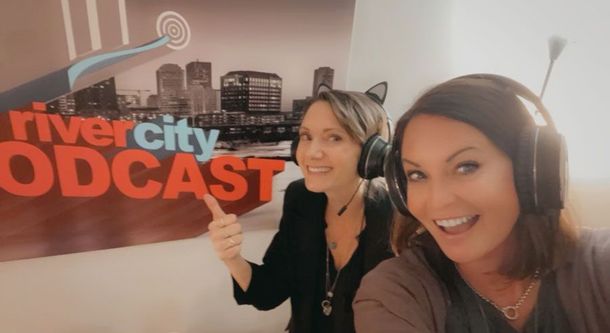 Two women smiling into the camera, wearing headphones, in front of a 