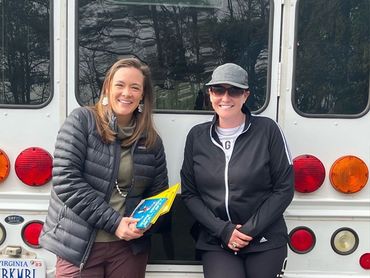 Two women smiling by a white bus. One holds a book. Trees in the background.