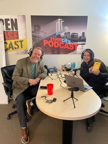 Two people in a podcast studio smiling. Man in blazer, woman with headphones, holding book, at a table with mics.