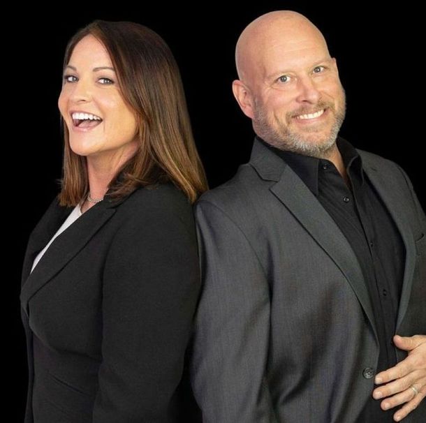 Woman and man smiling, standing back-to-back. Both wear suit jackets, black background.