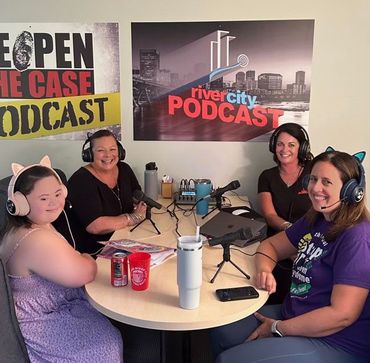 Four women recording a podcast, seated around a table with microphones and headphones, in front of podcast posters.