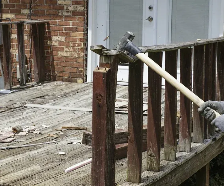 Person using a sledgehammer to demolish a wooden deck railing; exterior setting.