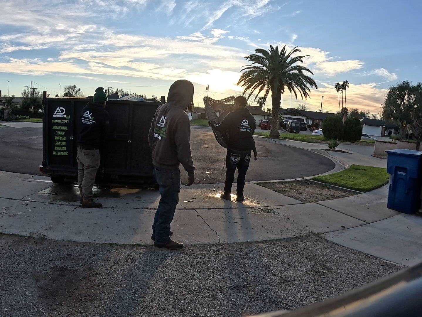 A&D Team near a dumpster under a sunset sky. Two appear to be moving it, while the other watches.