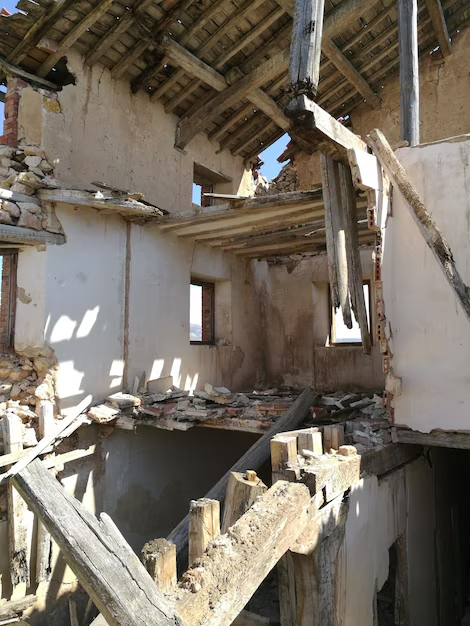 Interior of a dilapidated building with exposed wooden beams, crumbling walls, and two small windows.