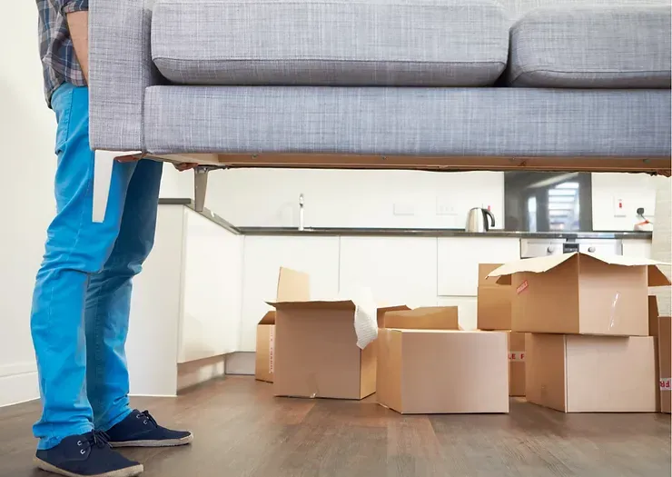 Person lifting a gray couch in a room with cardboard boxes.