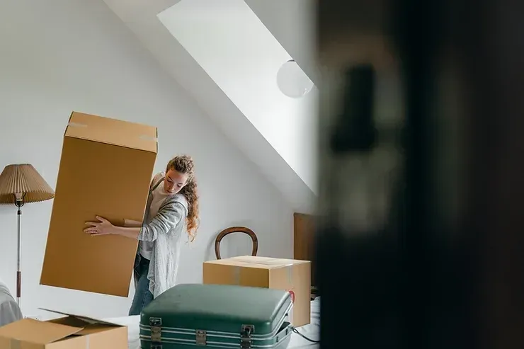 Woman lifting a large cardboard box inside a room with other boxes, possibly moving.