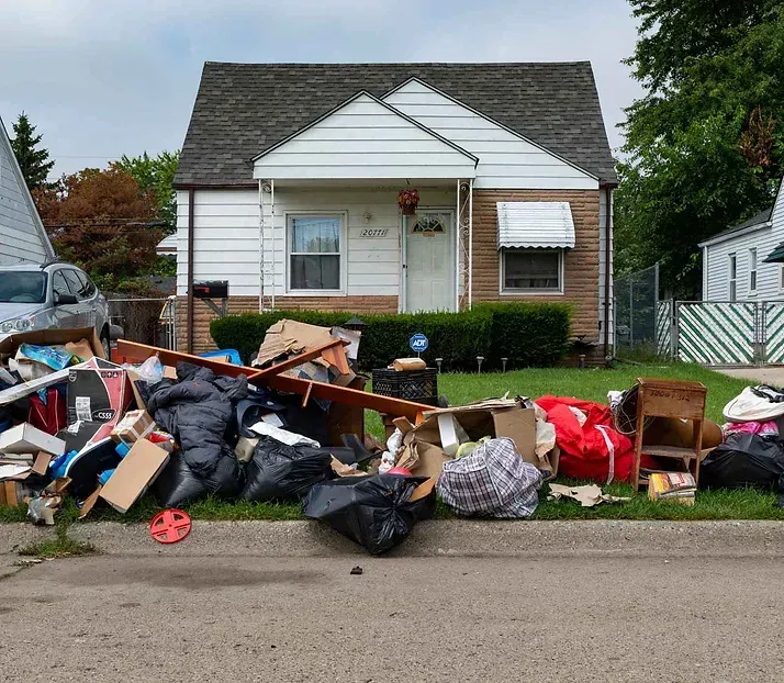 Pile of trash on curb in front of a small white house with a brown roof and a white awning.