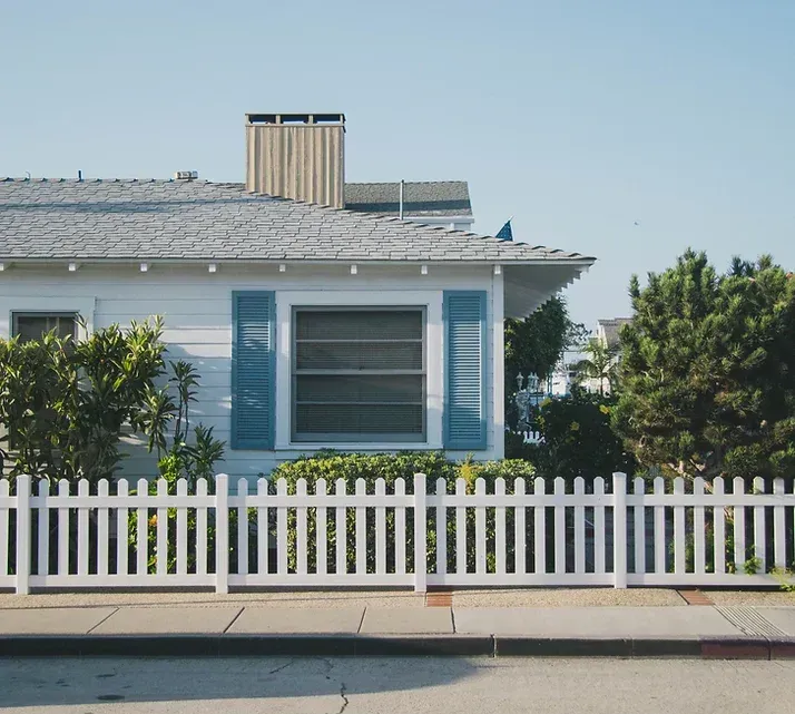 White house with blue shutters, white picket fence, and green bushes under a clear blue sky.
