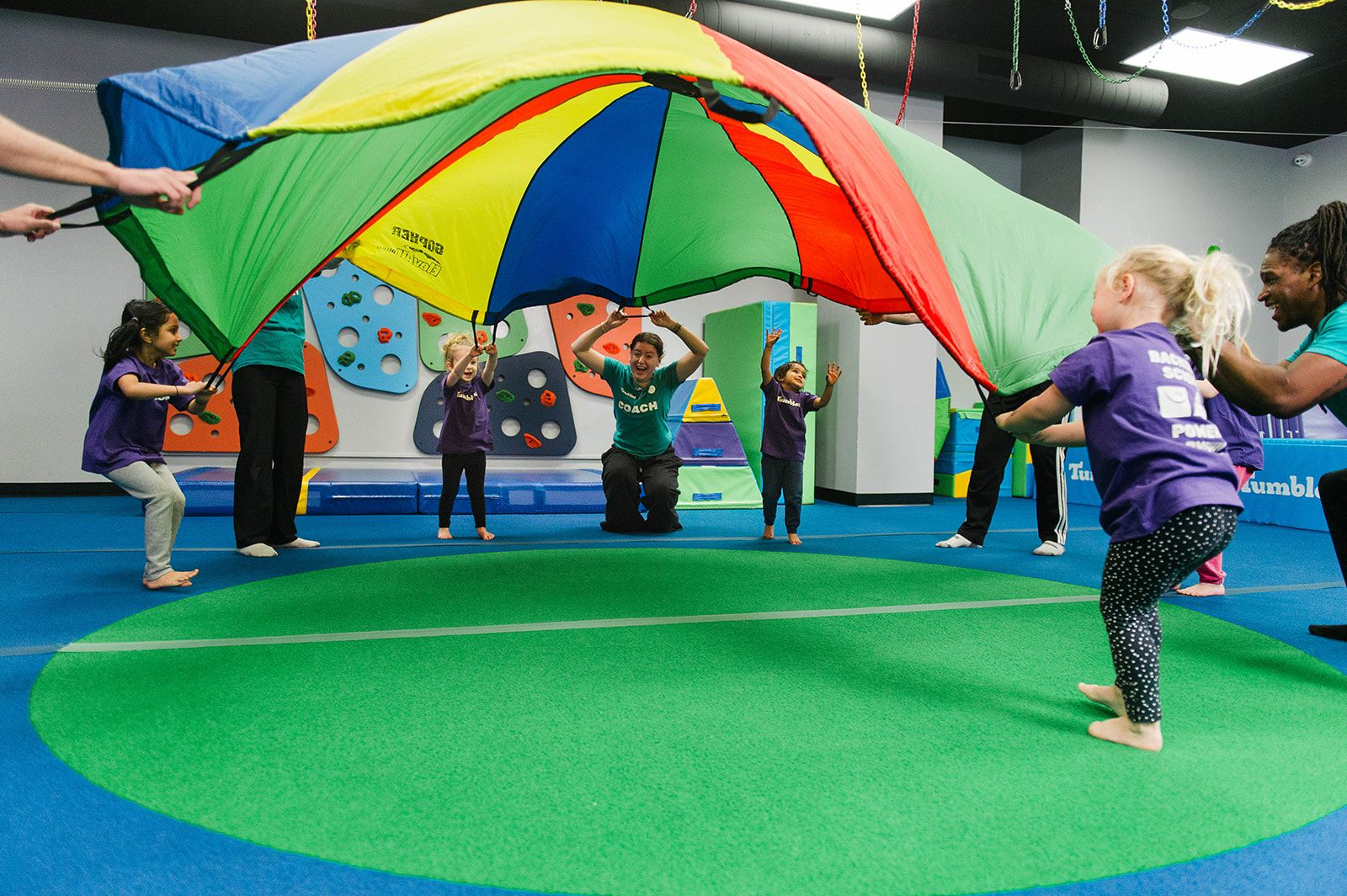 Children in a gymnasium gather around a green mat, with a teacher, interacting and observing.