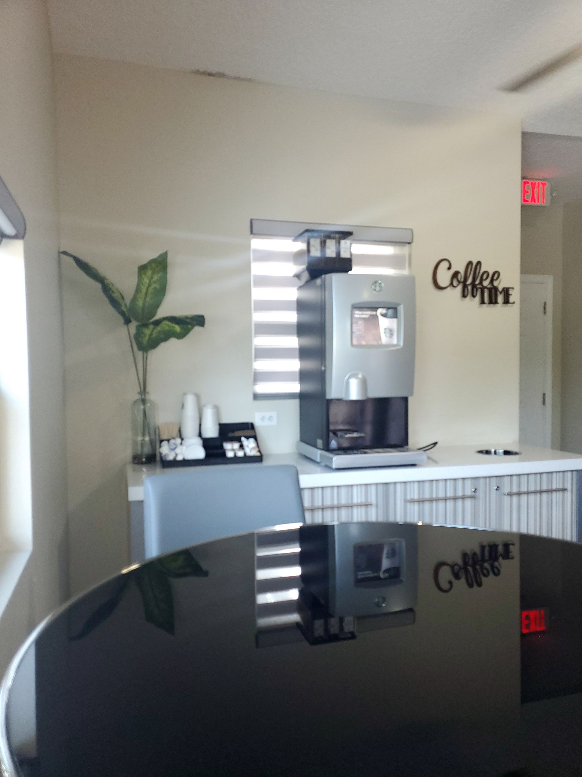 Coffee station with a coffee machine, cups, and a plant. Bright, with a reflective table.