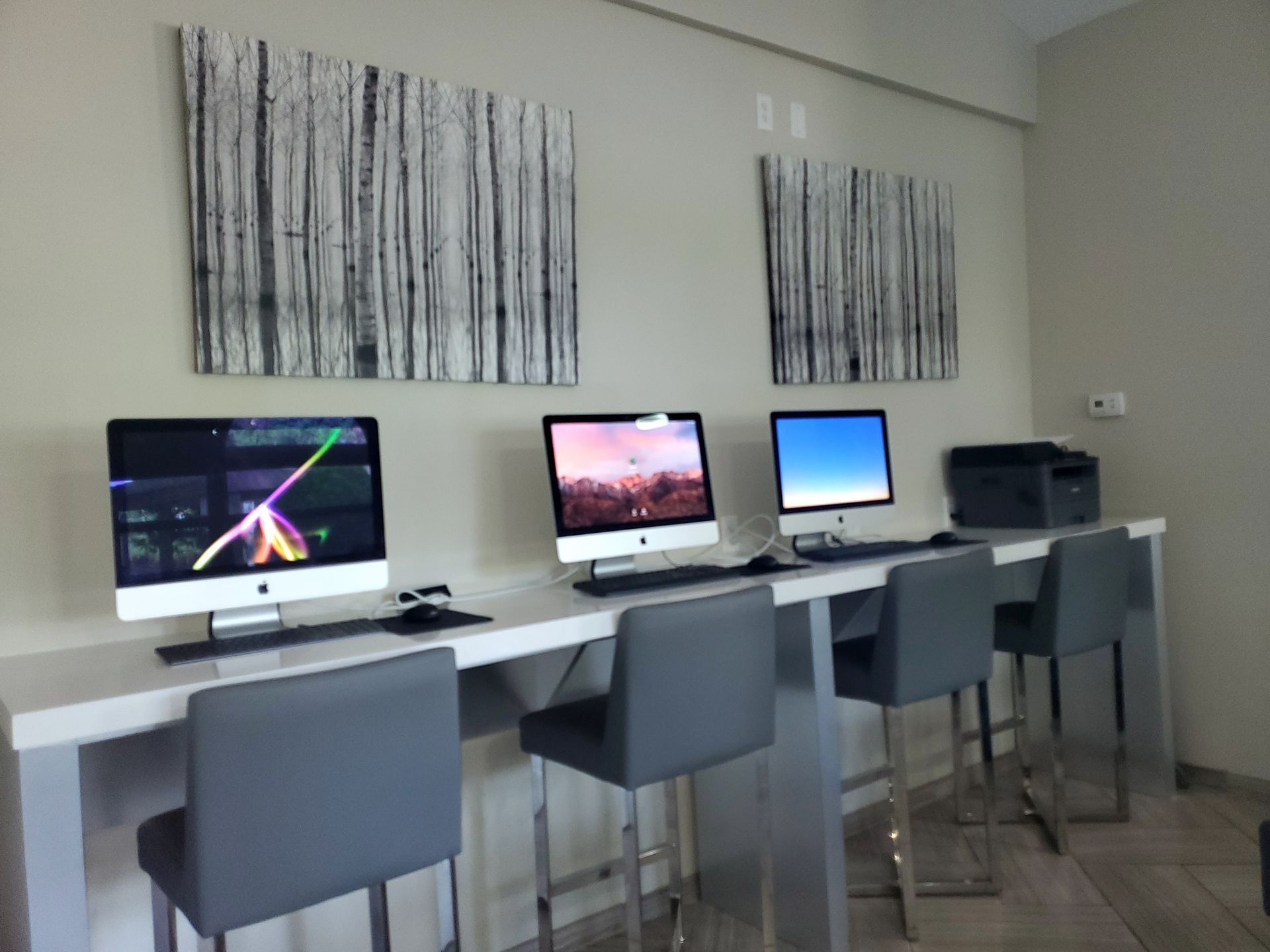 Computers on a white counter with gray chairs, printer, and artwork; a public workspace.