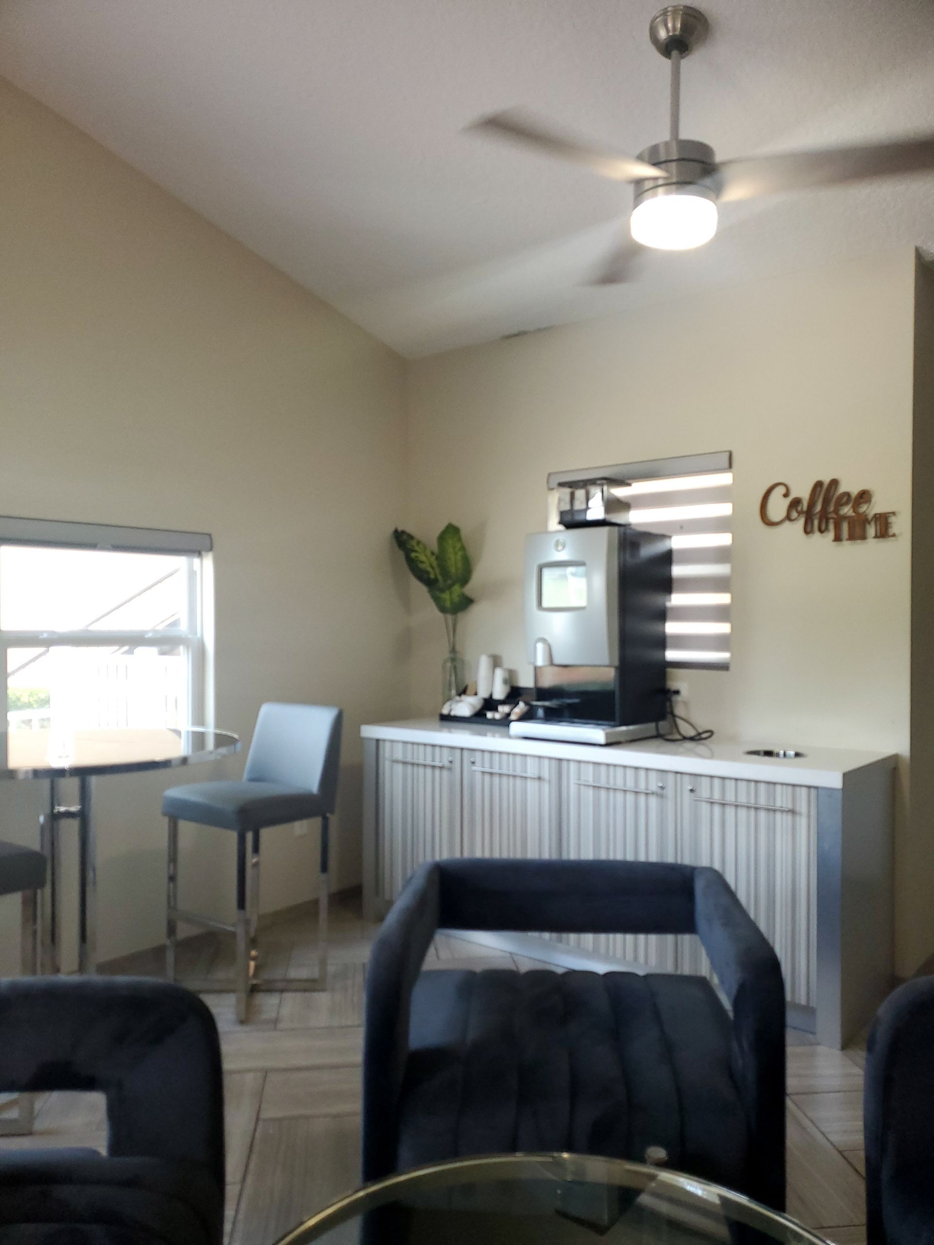 Coffee station with a machine, counter, and chairs in a room with a window and fan.