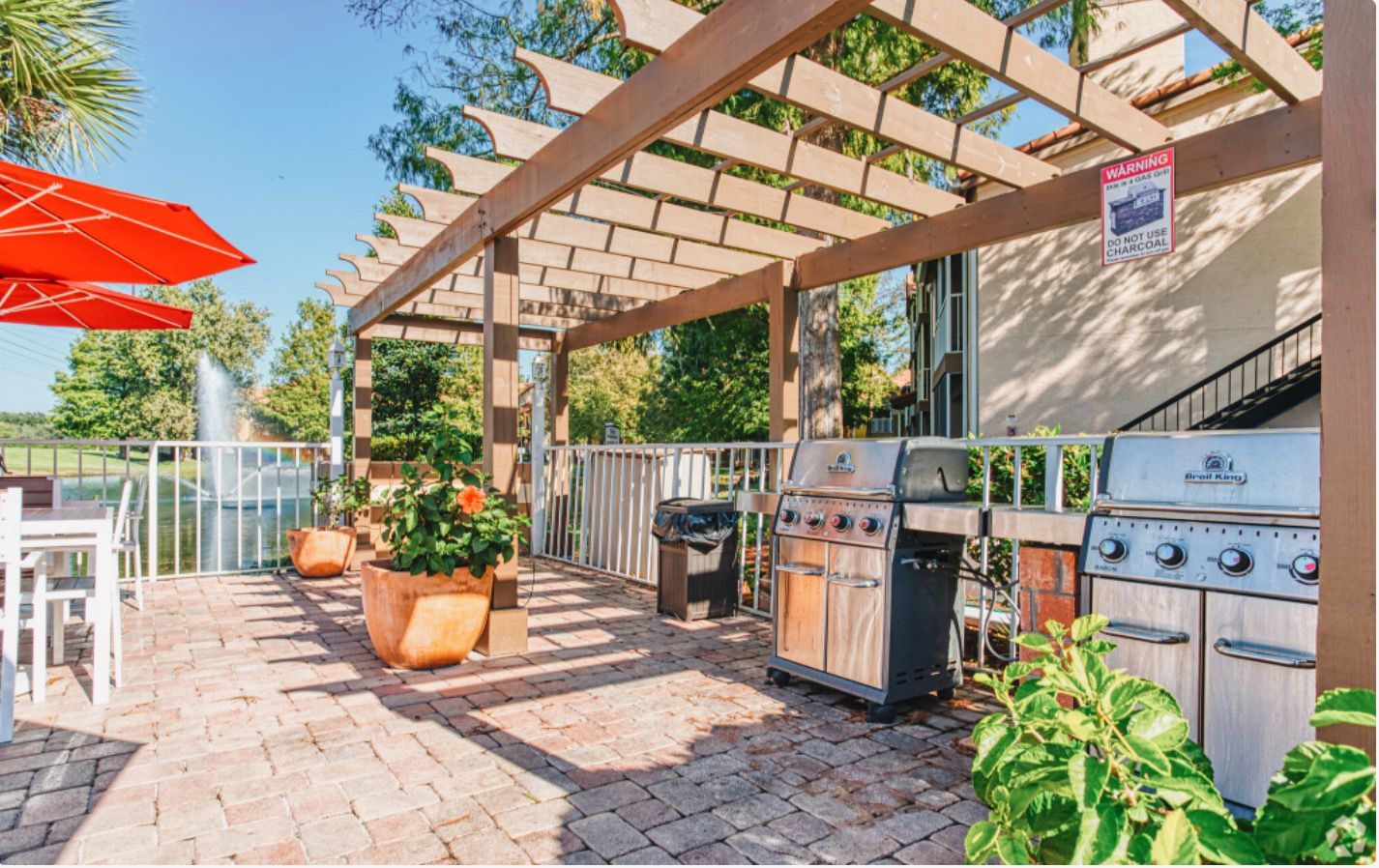 Outdoor grilling area with two grills under a wooden pergola, brick patio, fountain in background.