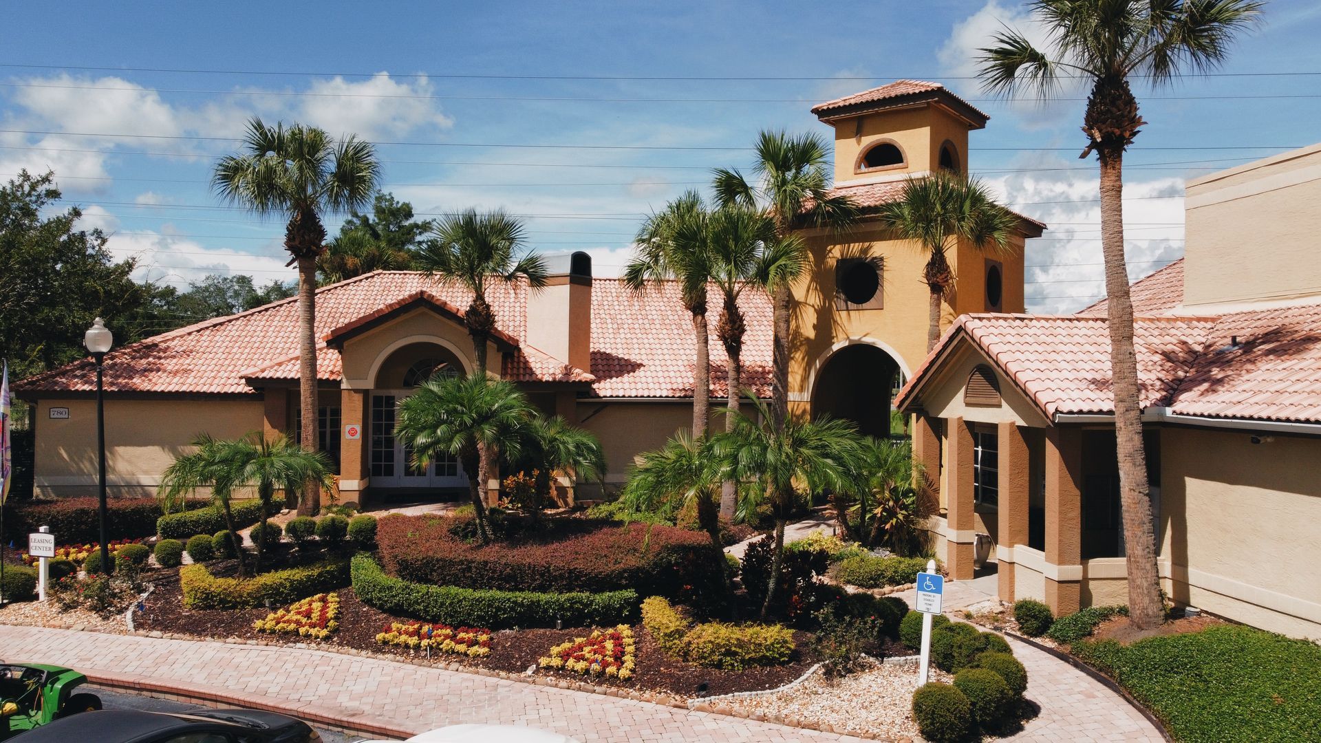 A tan, tile-roofed building with a tower, lush landscaping, and palm trees under a blue sky.