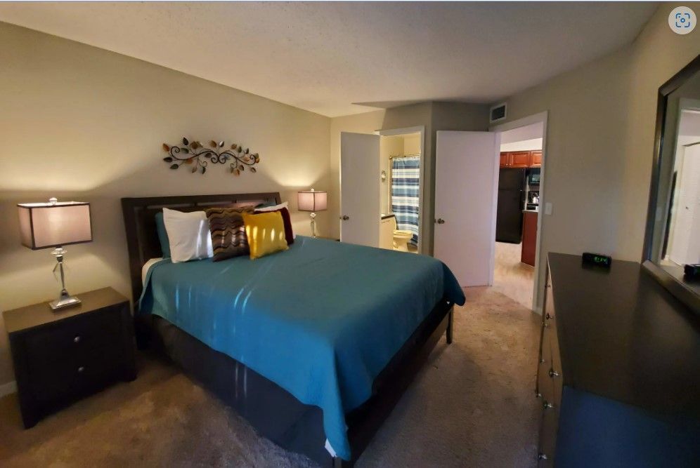 Bedroom with teal bedspread, nightstands, and dark dresser. Bathroom and kitchen visible from doorway.