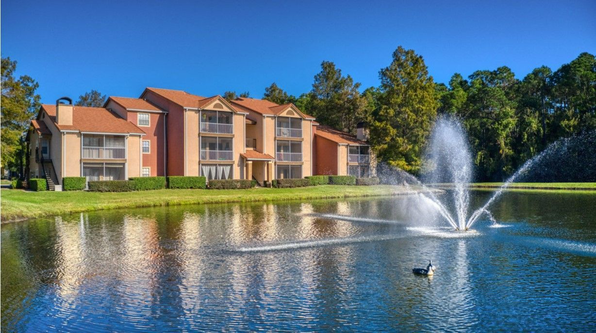 Apartment building beside a pond with a fountain. Blue sky, green trees, and a lone goose.