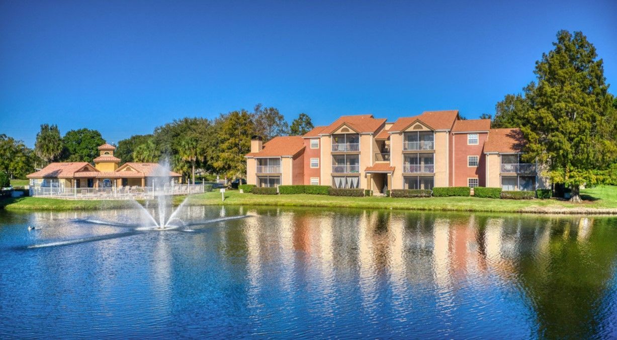 Apartment buildings with balconies beside a lake with a fountain under a blue sky.