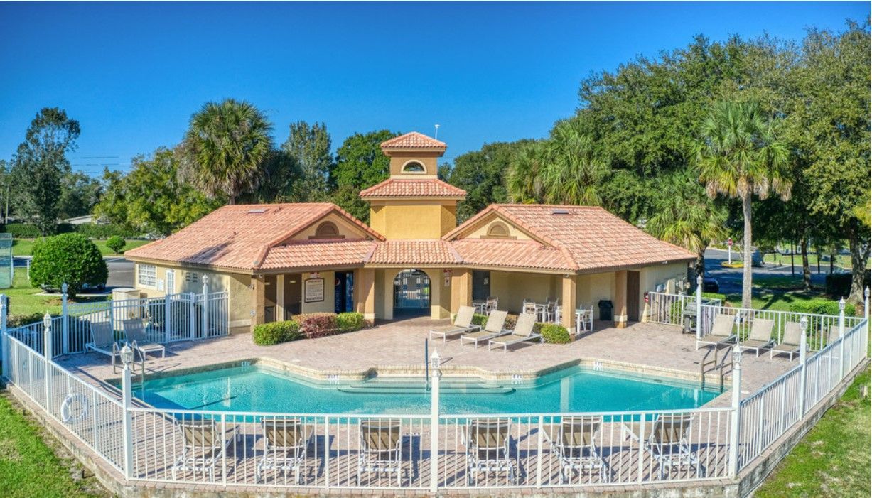 Swimming pool with beige building, lounge chairs, and fencing, set against greenery and a blue sky.