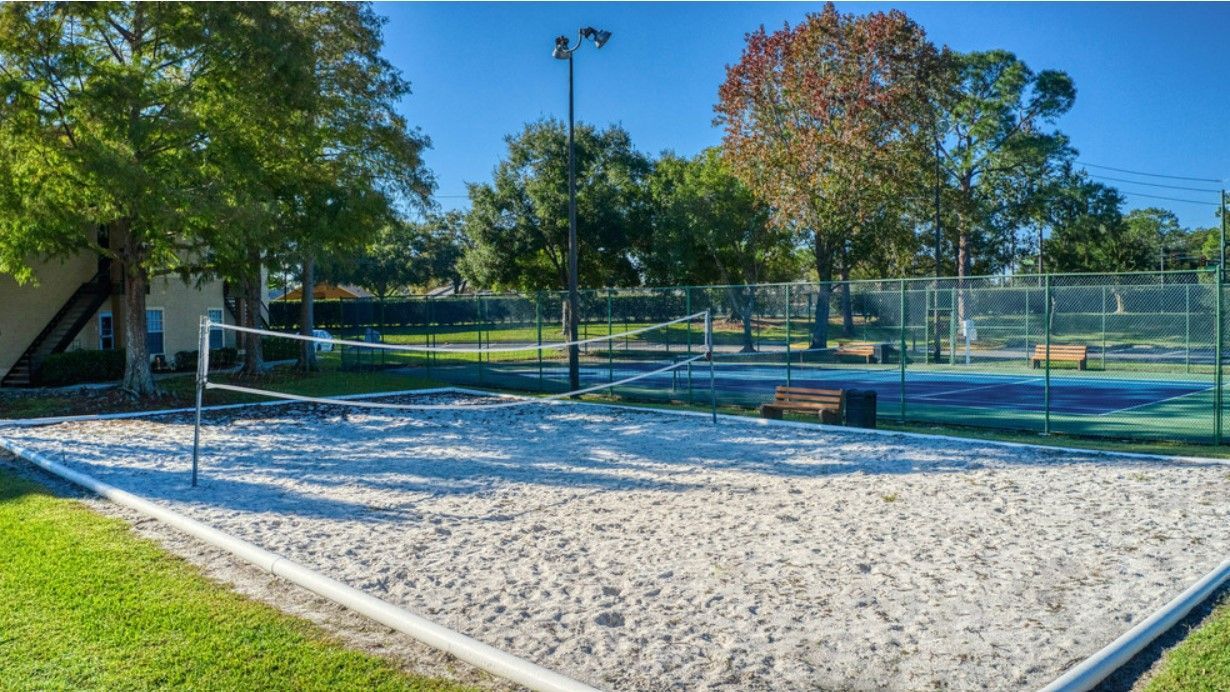 Sand volleyball court with net, trees, and tennis court in background.