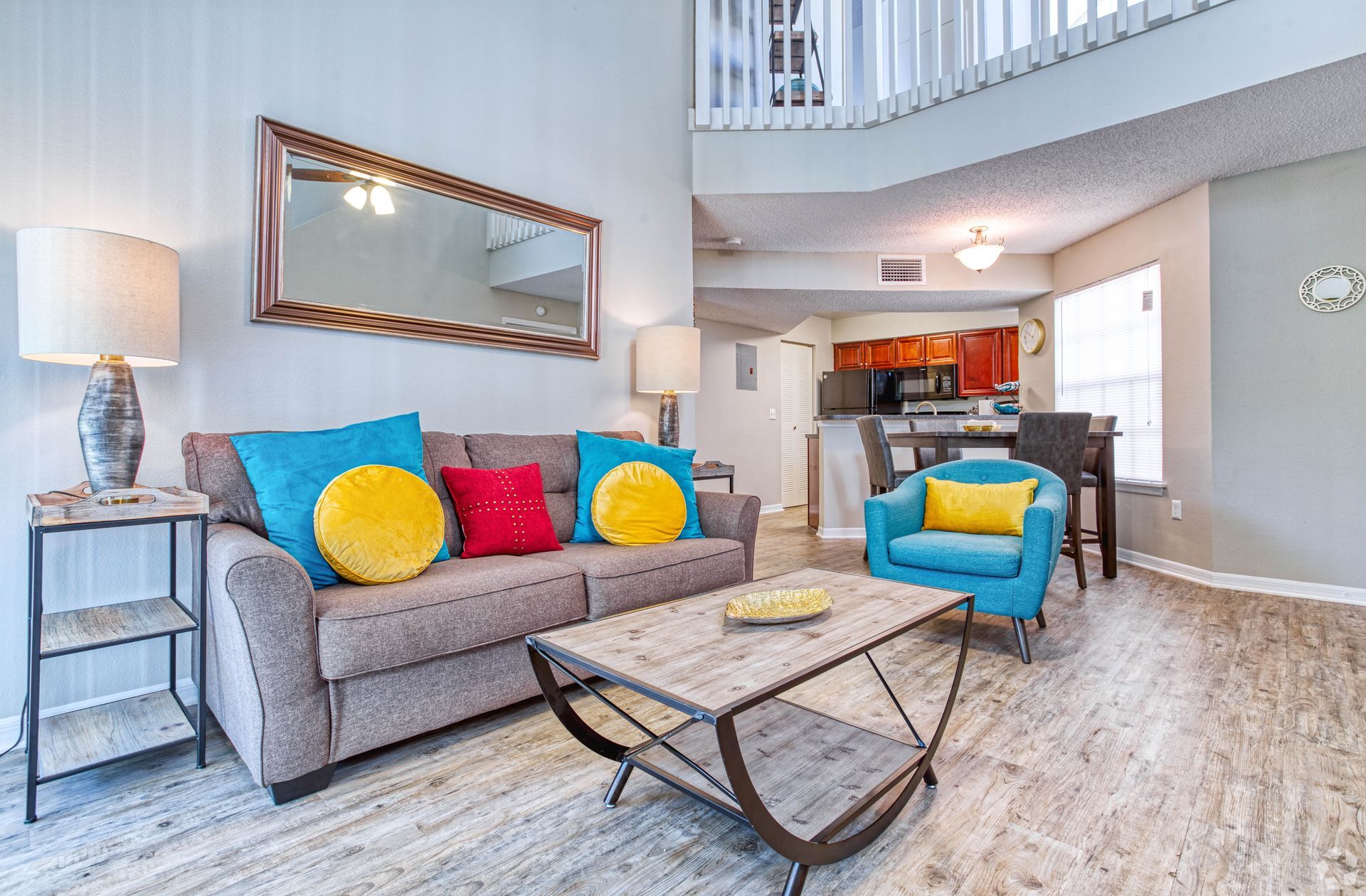 Living room with grey sofa, blue armchair, and colorful pillows. Light flooring, kitchen in background.