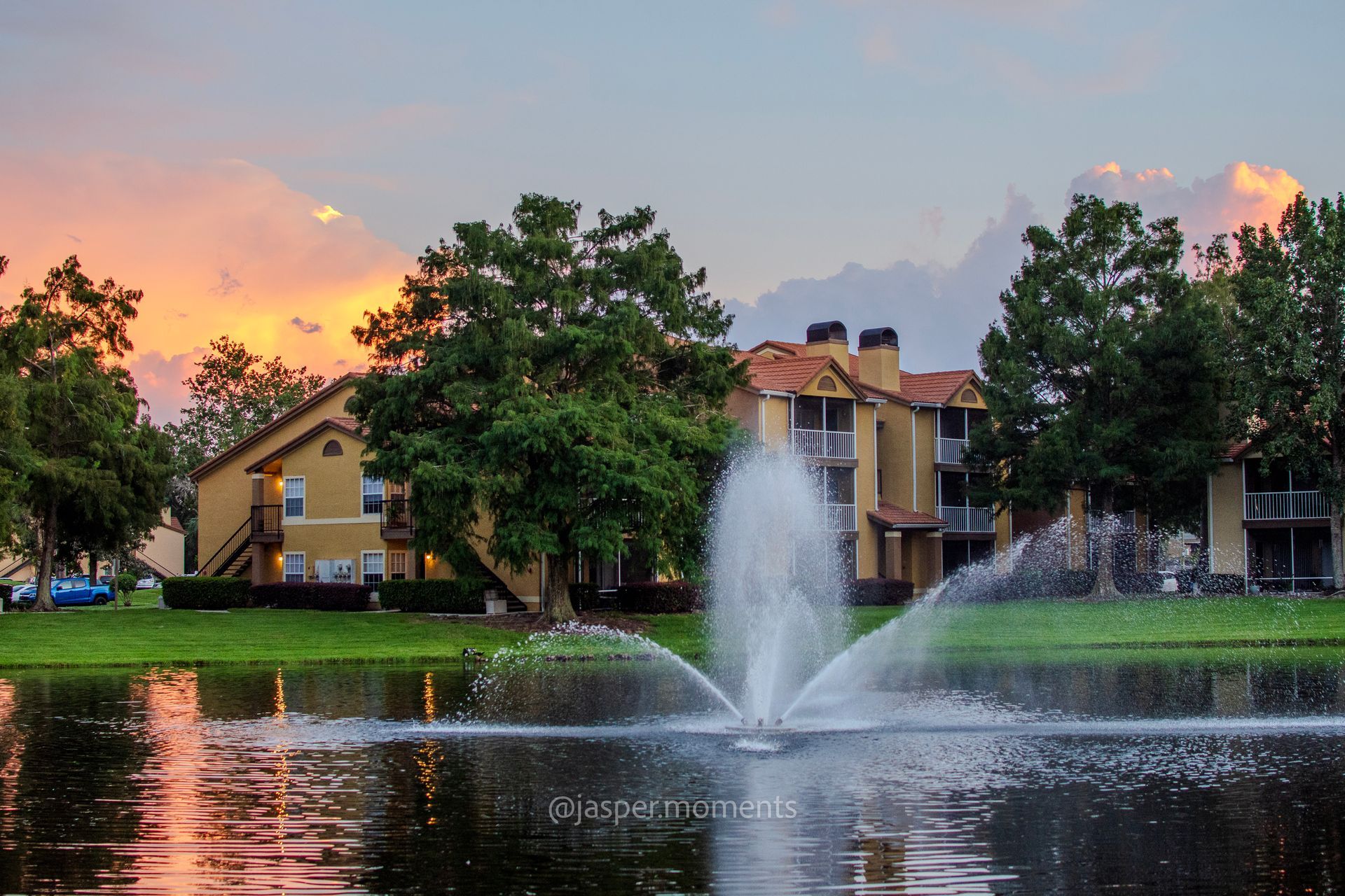 Fountain sprays water into a lake with apartment buildings and sunset sky backdrop.