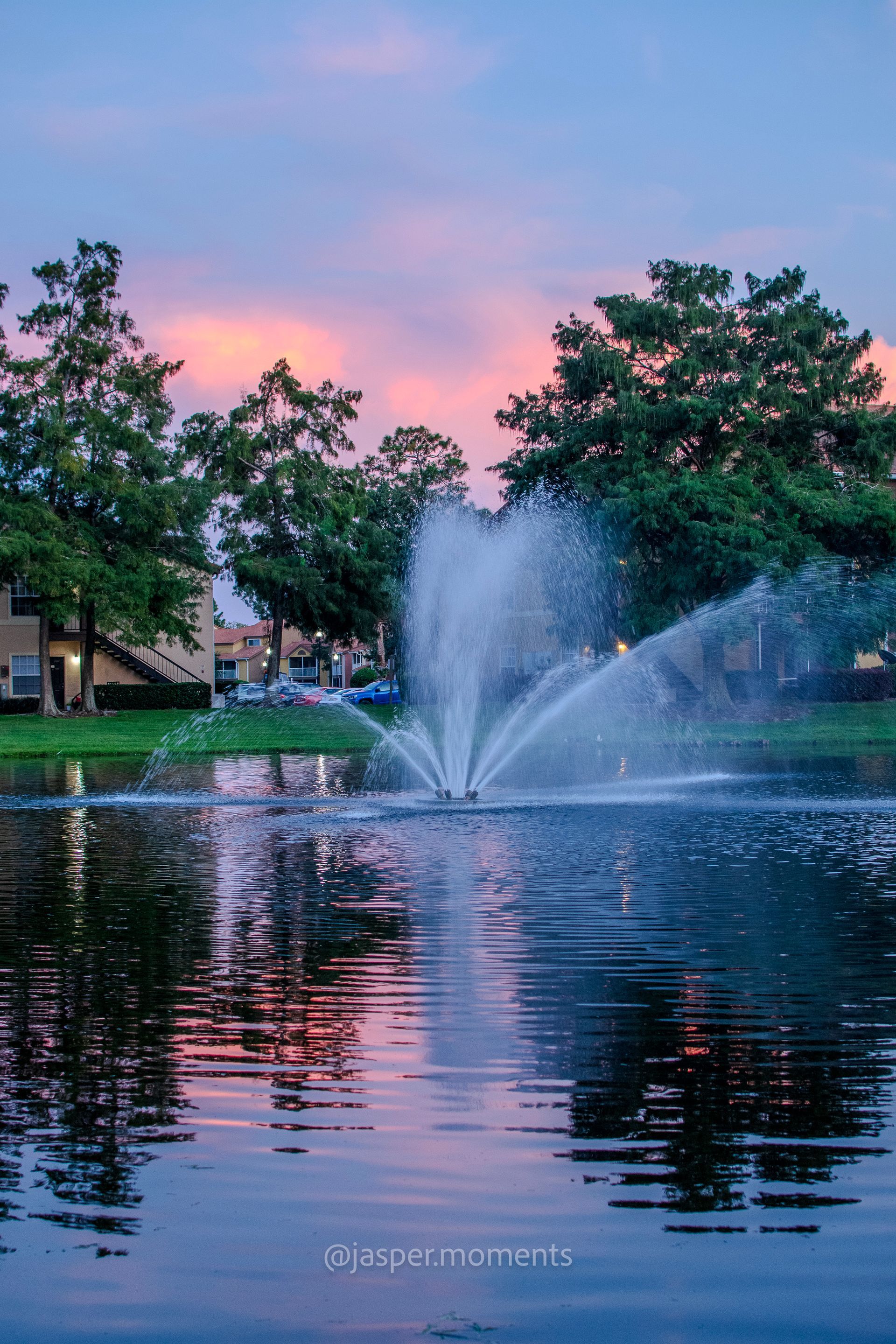 Fountain spraying water in a lake reflecting a pink and blue sunset, with trees and buildings in the background.
