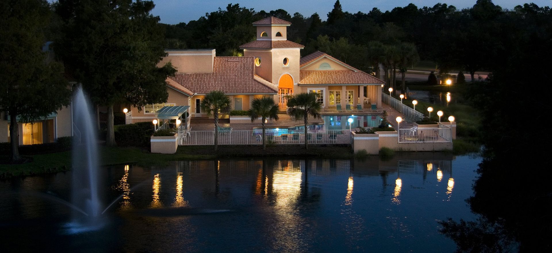 A building with a tower, lights, and a pool by a lake at dusk; fountain in front.