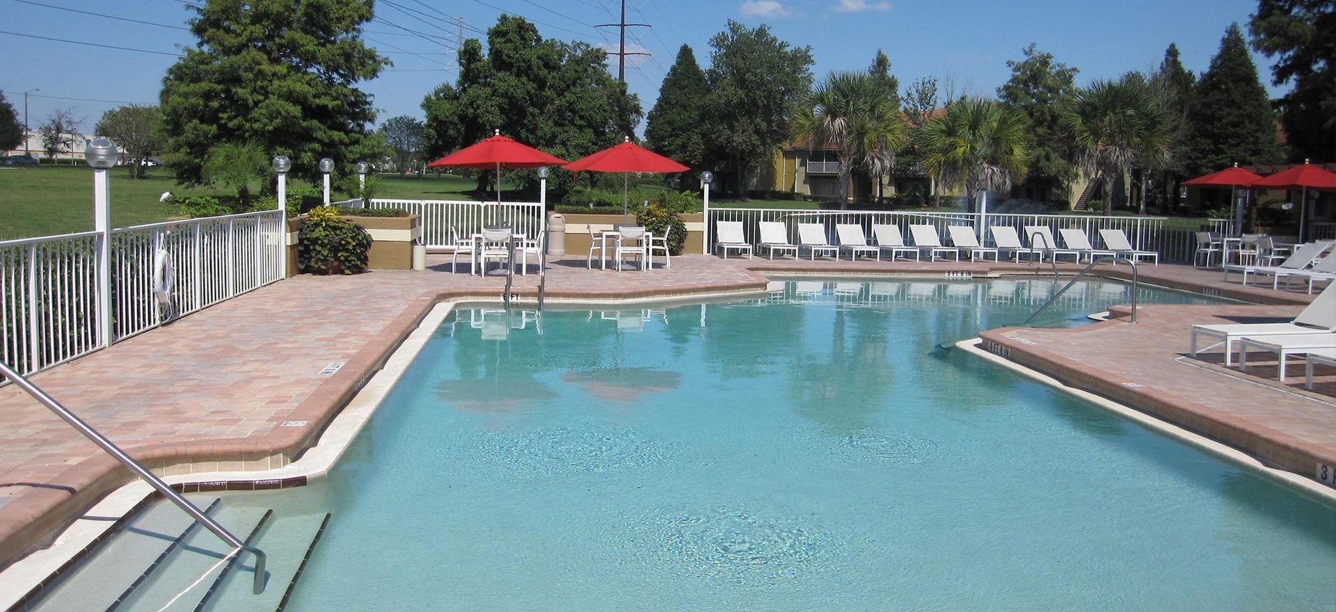 Swimming pool with lounge chairs, red umbrellas, and trees on a sunny day.