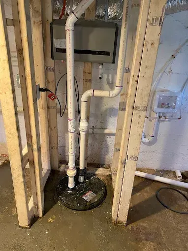 Basement sump pump system with white PVC pipes, electrical box, and wooden framing.