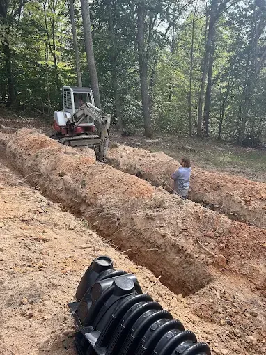 An excavator digs trenches in a dirt yard near a wooded area. A person stands nearby.