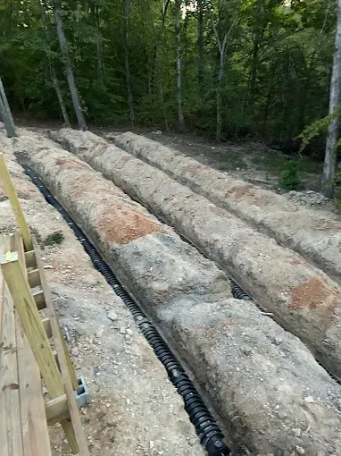 Trenches dug in earth for drainage system installation near a wooden deck and trees.