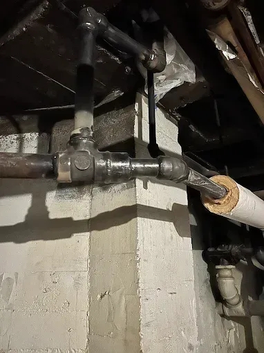 Black pipes and fittings in a basement, close to a concrete pillar. One pipe is insulated.