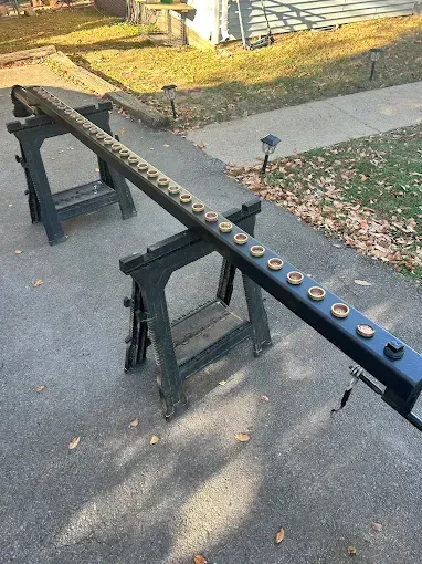 Black, long object with many golden holes on sawhorses outside on asphalt.