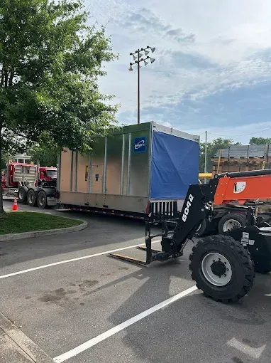 Large metal container on a flatbed trailer being moved by a forklift in a parking lot. A red semi-truck is visible.
