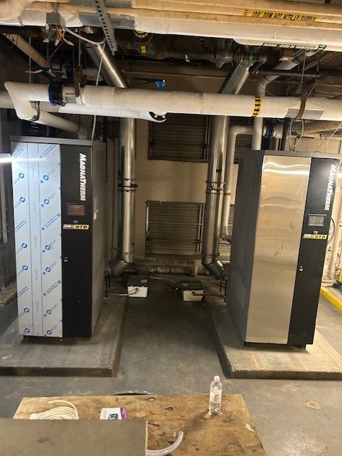 Two large, black and silver industrial machines in a utility room, with pipes and insulation overhead.