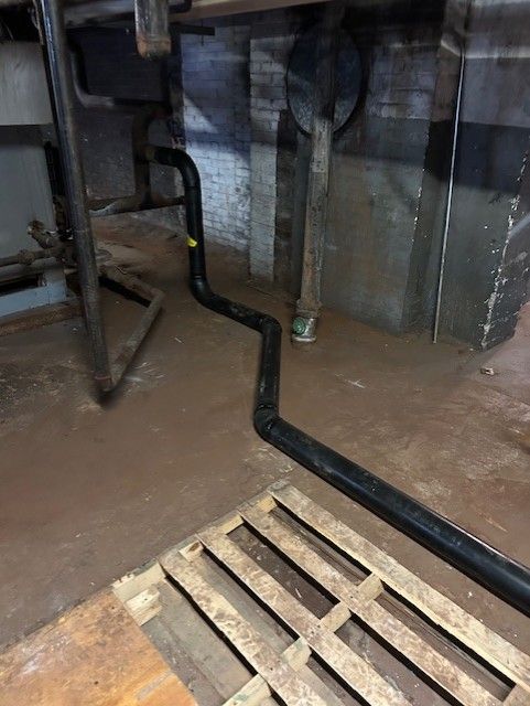 Black pipe zig-zags across a brown, damp floor with a wooden grate, running toward a brick wall.