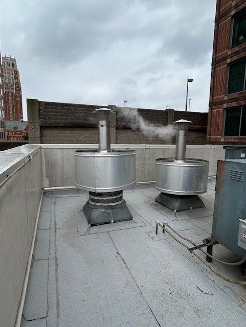 Two stainless steel exhaust vents on a rooftop, emitting white vapor. Gray, cloudy sky.
