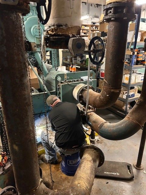 A person works on machinery in an industrial setting, surrounded by pipes and equipment.