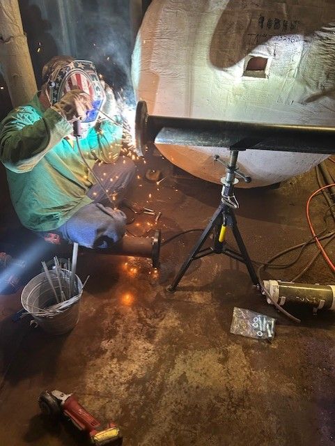 Welder wearing a mask, welding a pipe to a metal structure in a dim indoor setting, sparks flying.