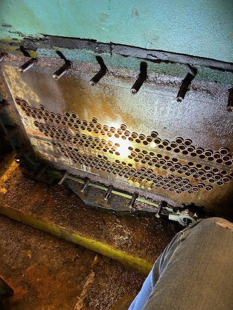 Close-up of a metal plate with numerous holes, possibly a boiler component, in a dim, industrial setting.