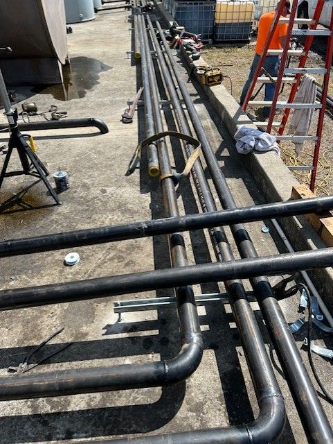 Black pipes laid out on concrete, with tools and a ladder, likely for construction.