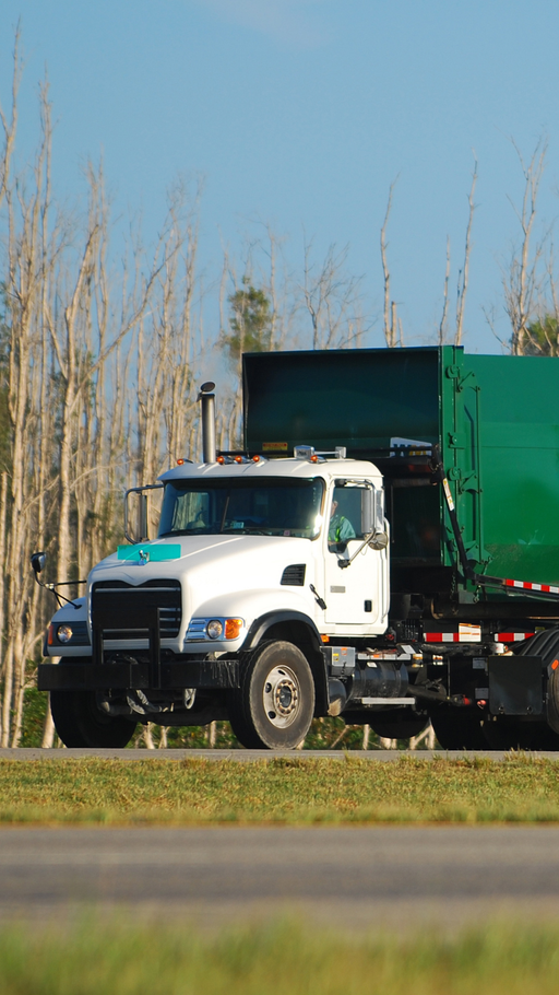 A white garbage truck is driving down the road next to a green garbage truck.