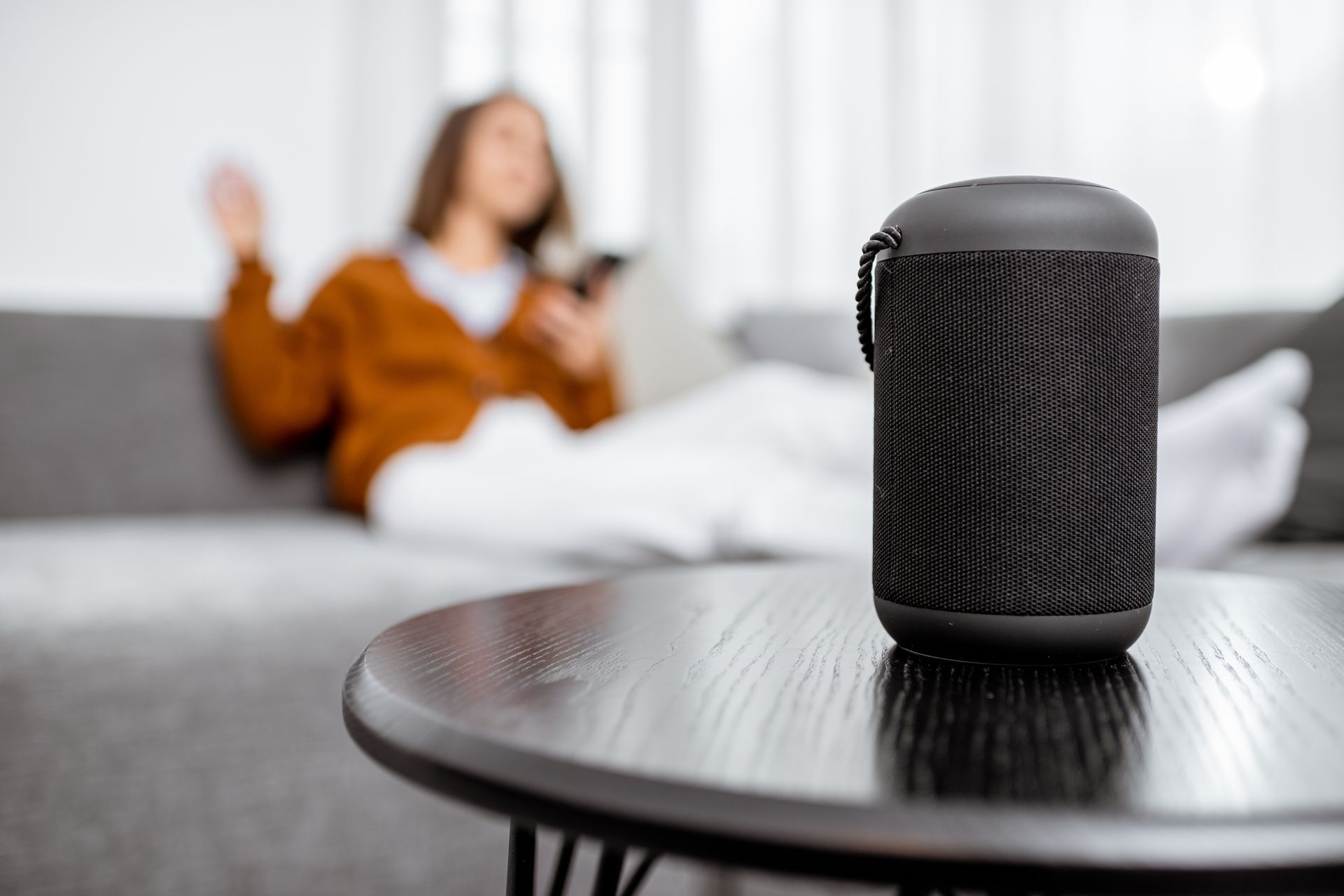 Black cylindrical speaker on a table; person relaxing on a sofa in the background.